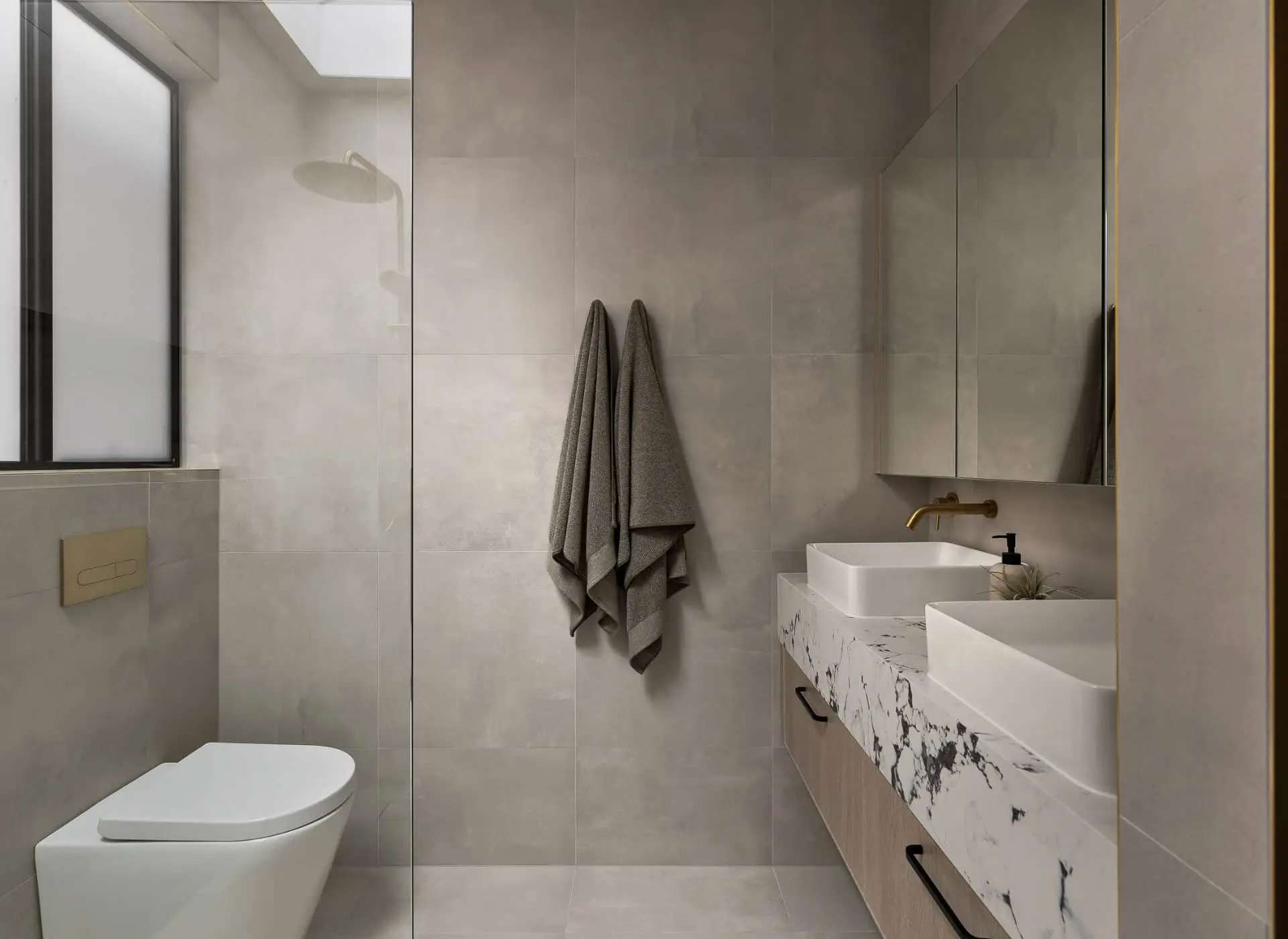 Modern bathroom with light gray tiles, white toilet, double vessel sinks on marble countertop, and gray towels hanging on wall.