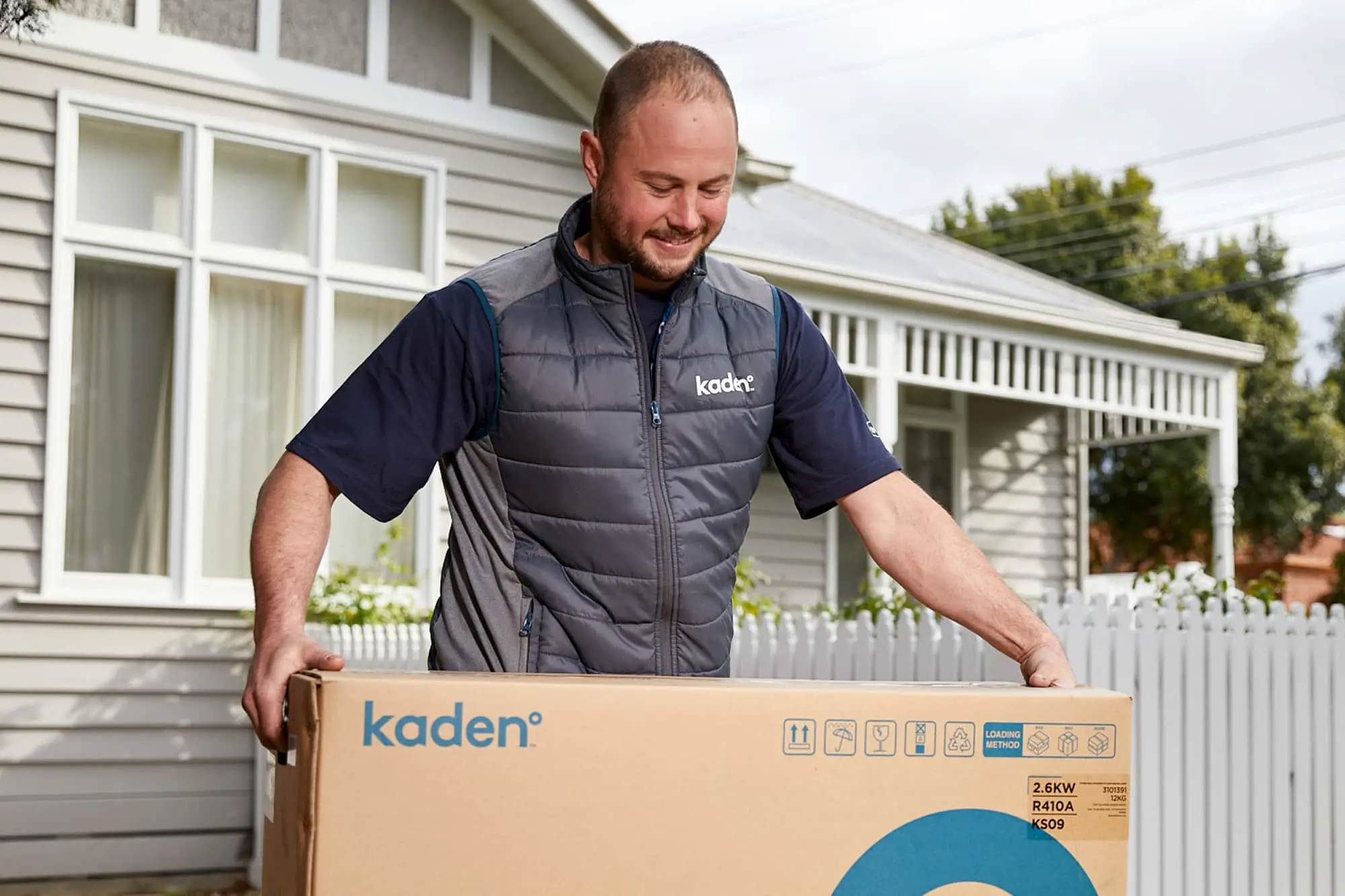 Person in gray Kaden vest delivering a boxed appliance outside a white house with picket fence.