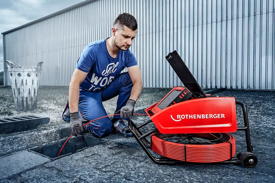 Person in blue workwear using a red Rothenberger drain cleaning machine outside a metal building.