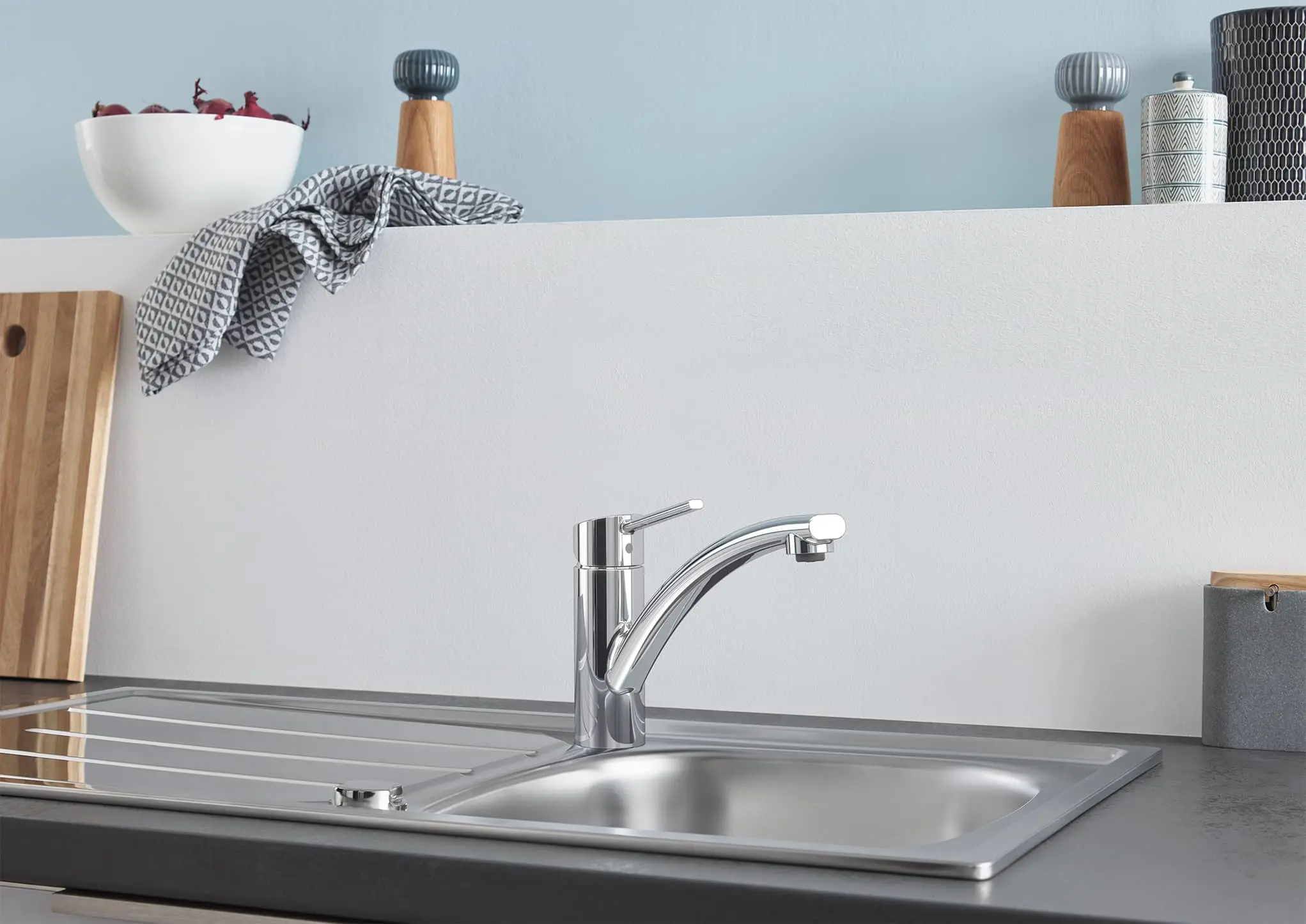 Modern kitchen sink with chrome faucet, white shelf holding red onions in bowl, pepper mills, and patterned kitchen towel.