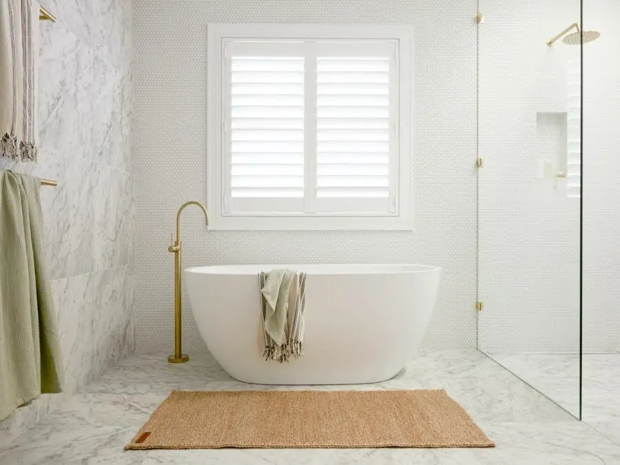 Modern white bathroom with freestanding tub, marble walls, window with shutters, gold fixtures, and jute rug.