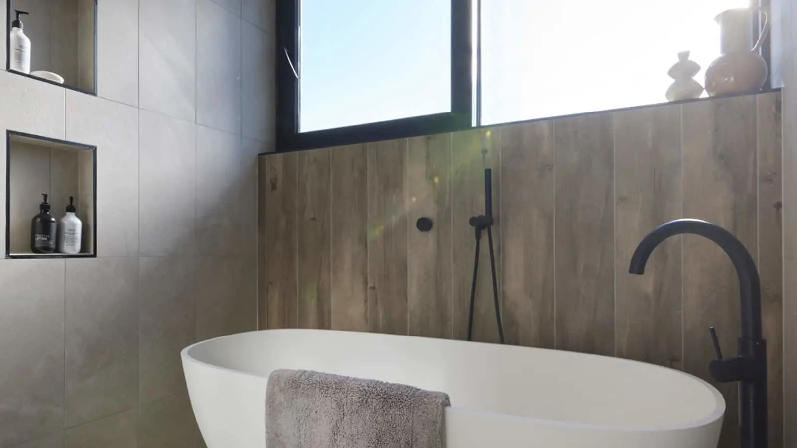 Modern bathroom with white freestanding tub, black fixtures, wood-look wall panels, and built-in shelving for toiletries.