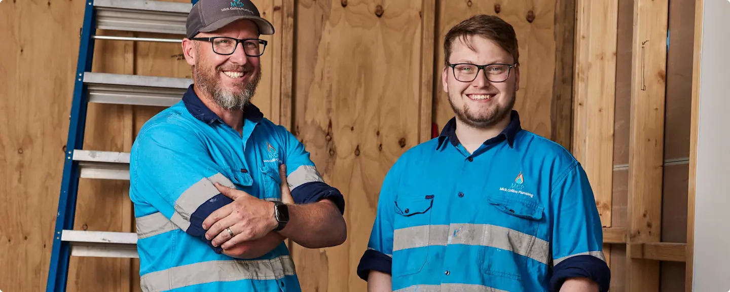 Two workers in blue safety uniforms with reflective stripes standing in front of wooden wall with ladder nearby.