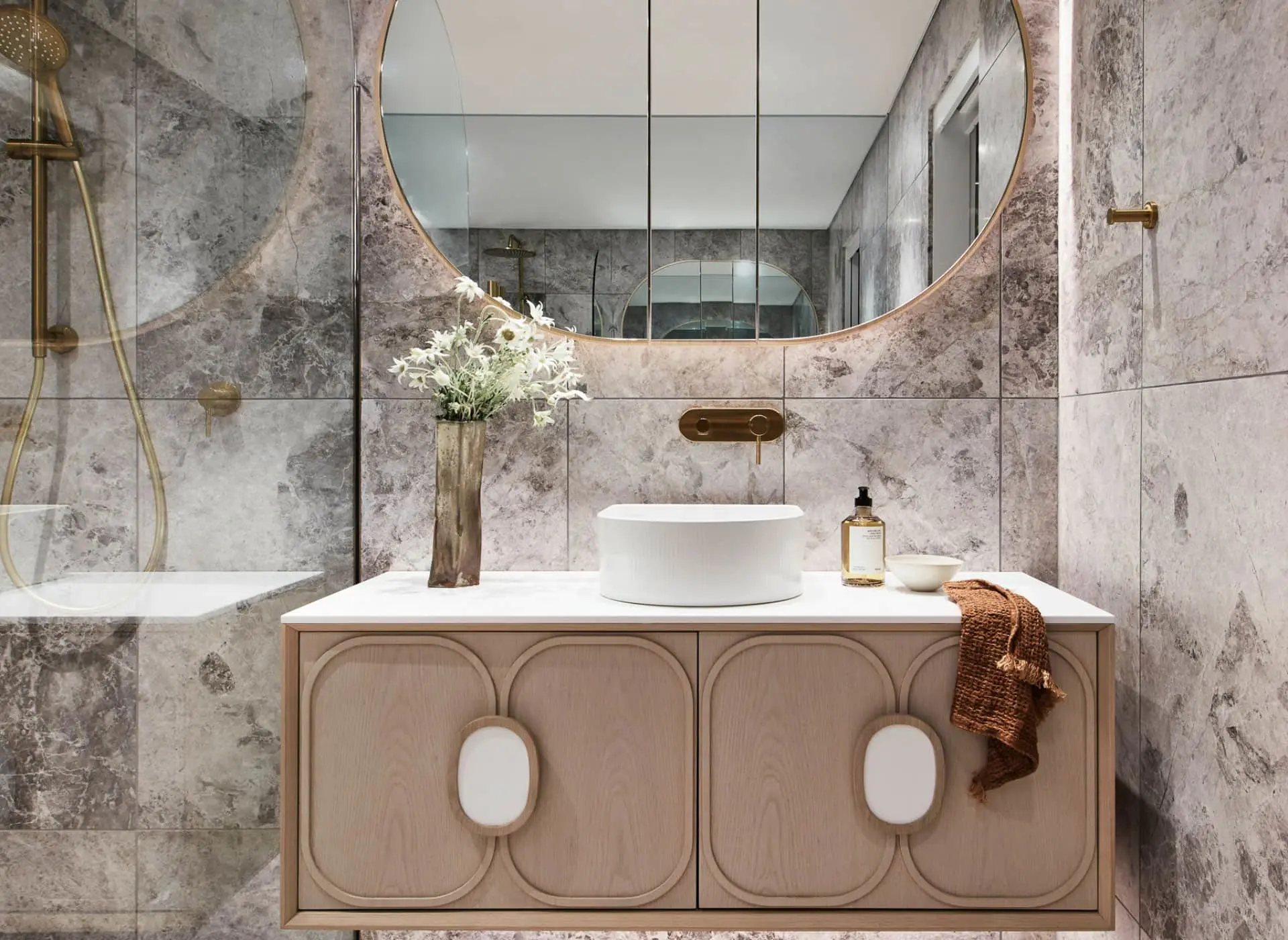 Modern bathroom with marble walls, round mirror, wooden vanity, white vessel sink, and gold fixtures.