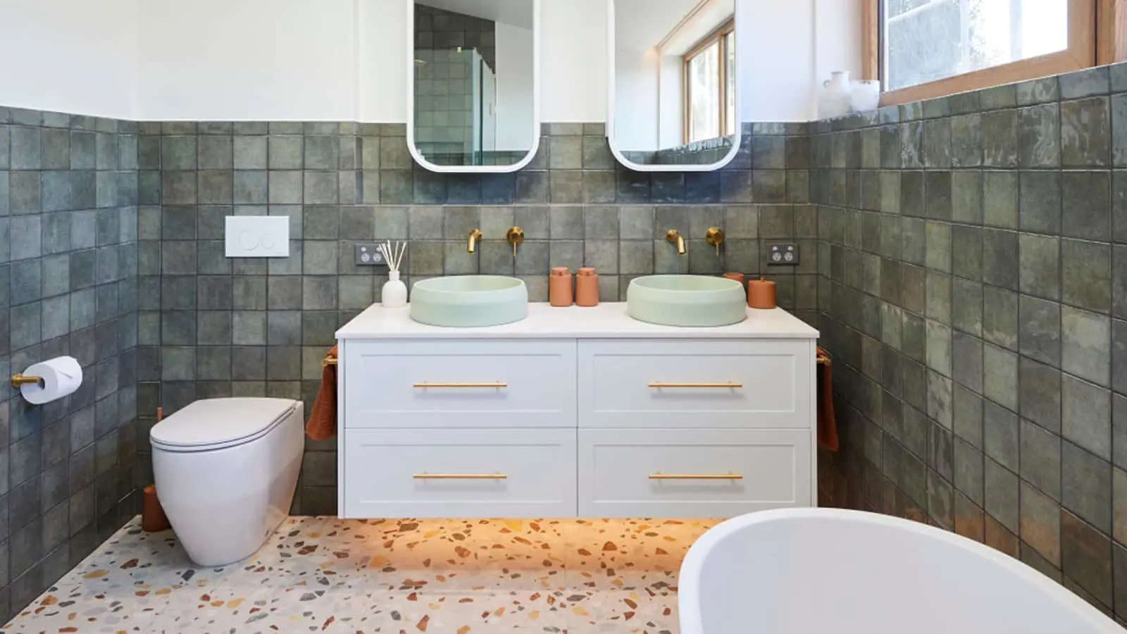 Modern bathroom with green tile walls, white vanity with dual mint sinks, gold fixtures, and terrazzo flooring.