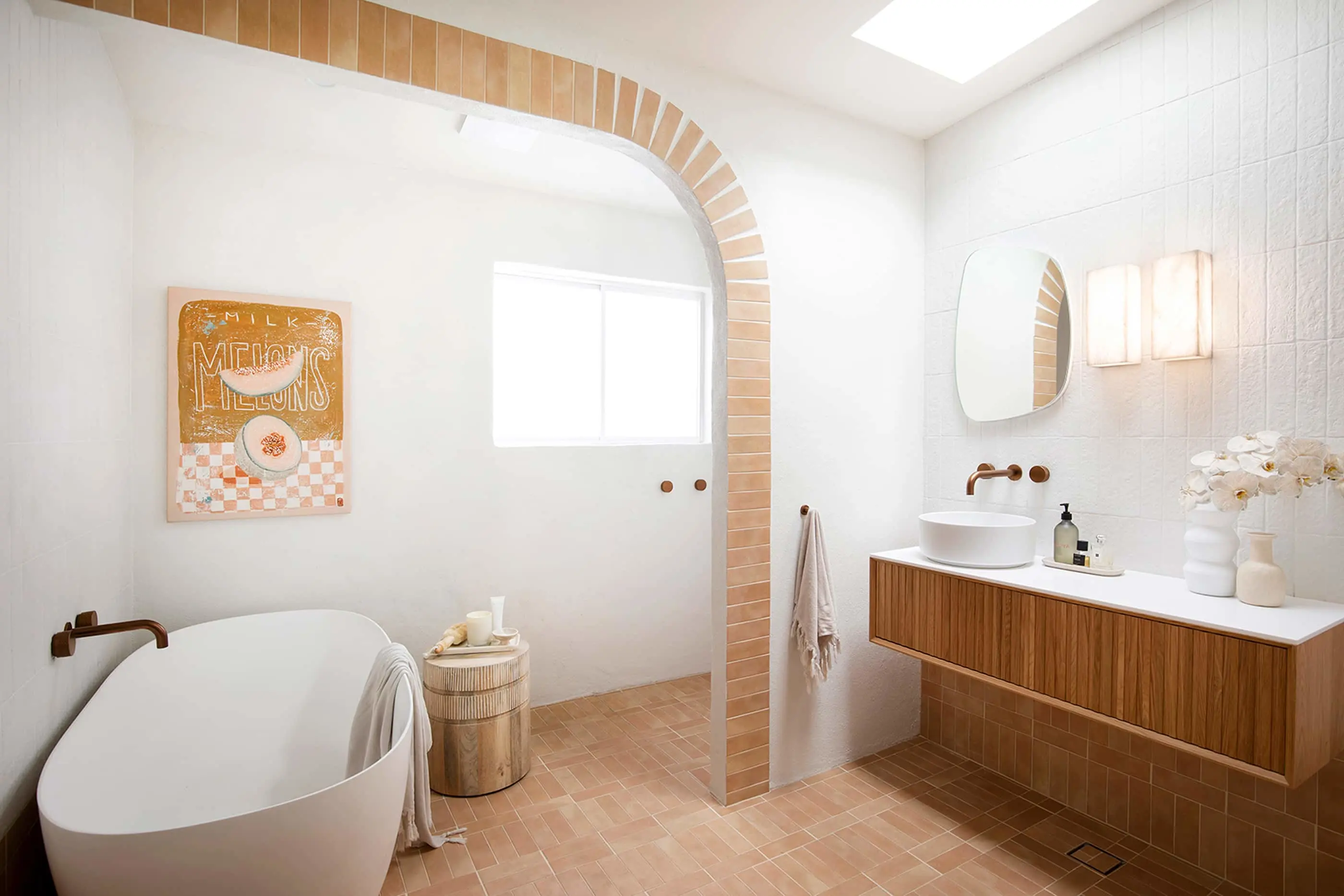 Modern bathroom with white freestanding tub, terracotta tile arch, wooden vanity, and orange artwork on white walls.