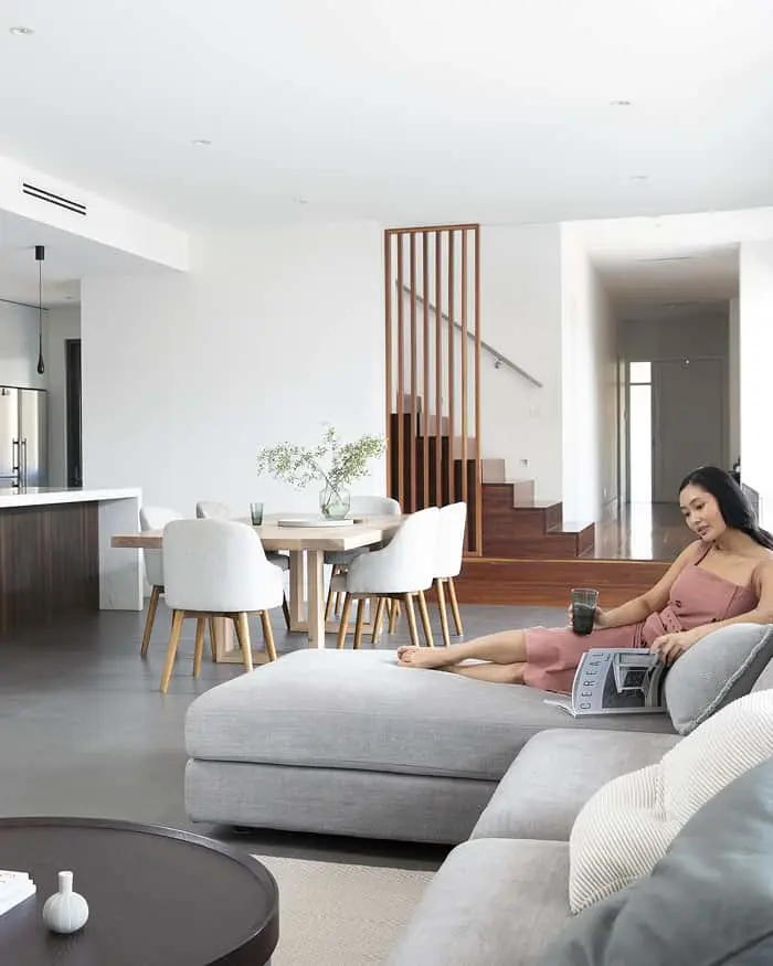 Modern open-concept living space with dining area, wooden staircase, and person relaxing on gray sectional sofa with magazine.