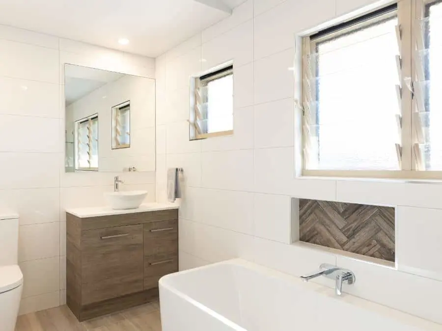 Modern white bathroom with wooden vanity, white bathtub, herringbone tile accent, and louvered windows.