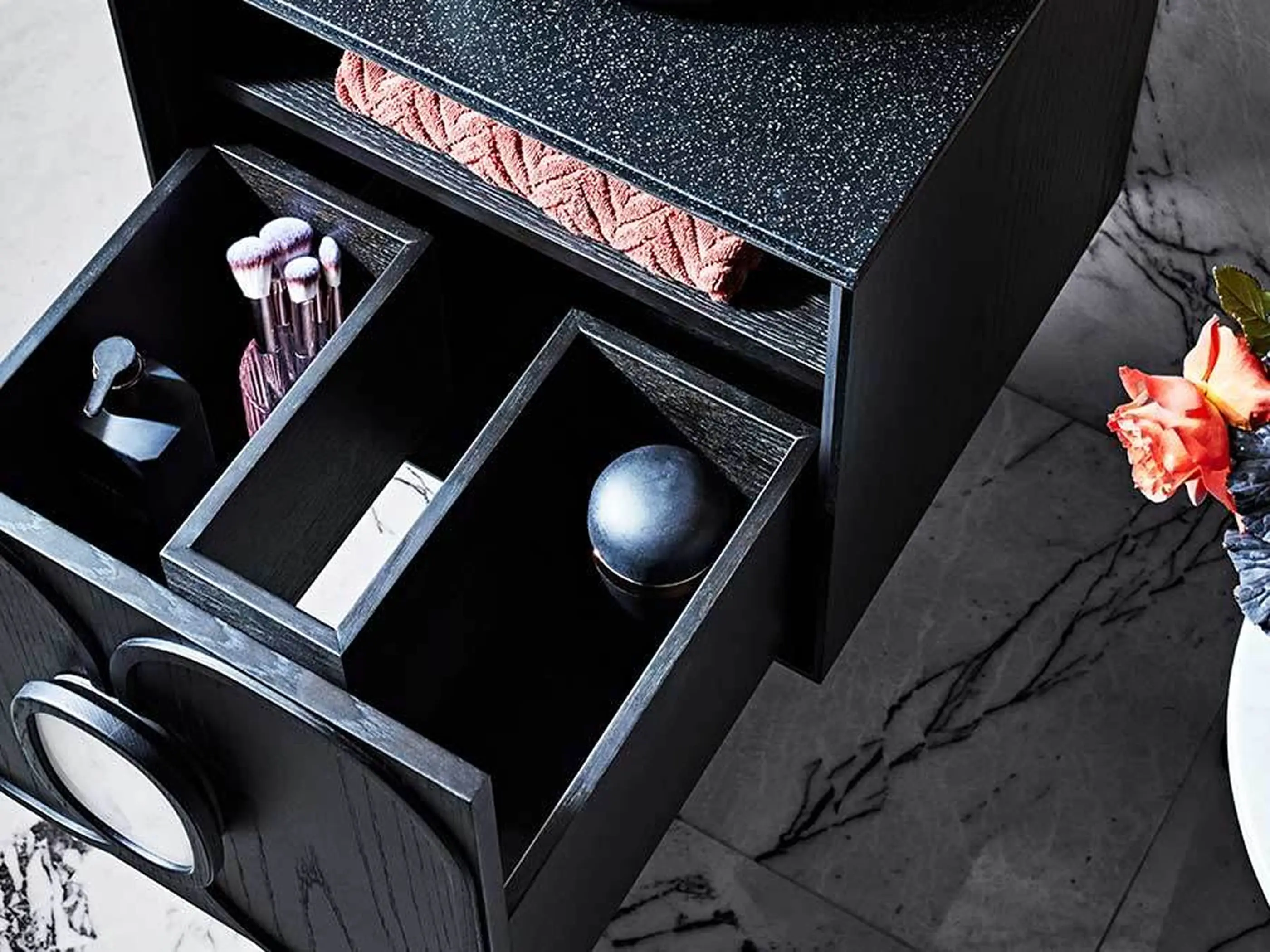 Black makeup organizer with open drawers containing makeup brushes, cosmetics, and a pink towel on marble countertop.