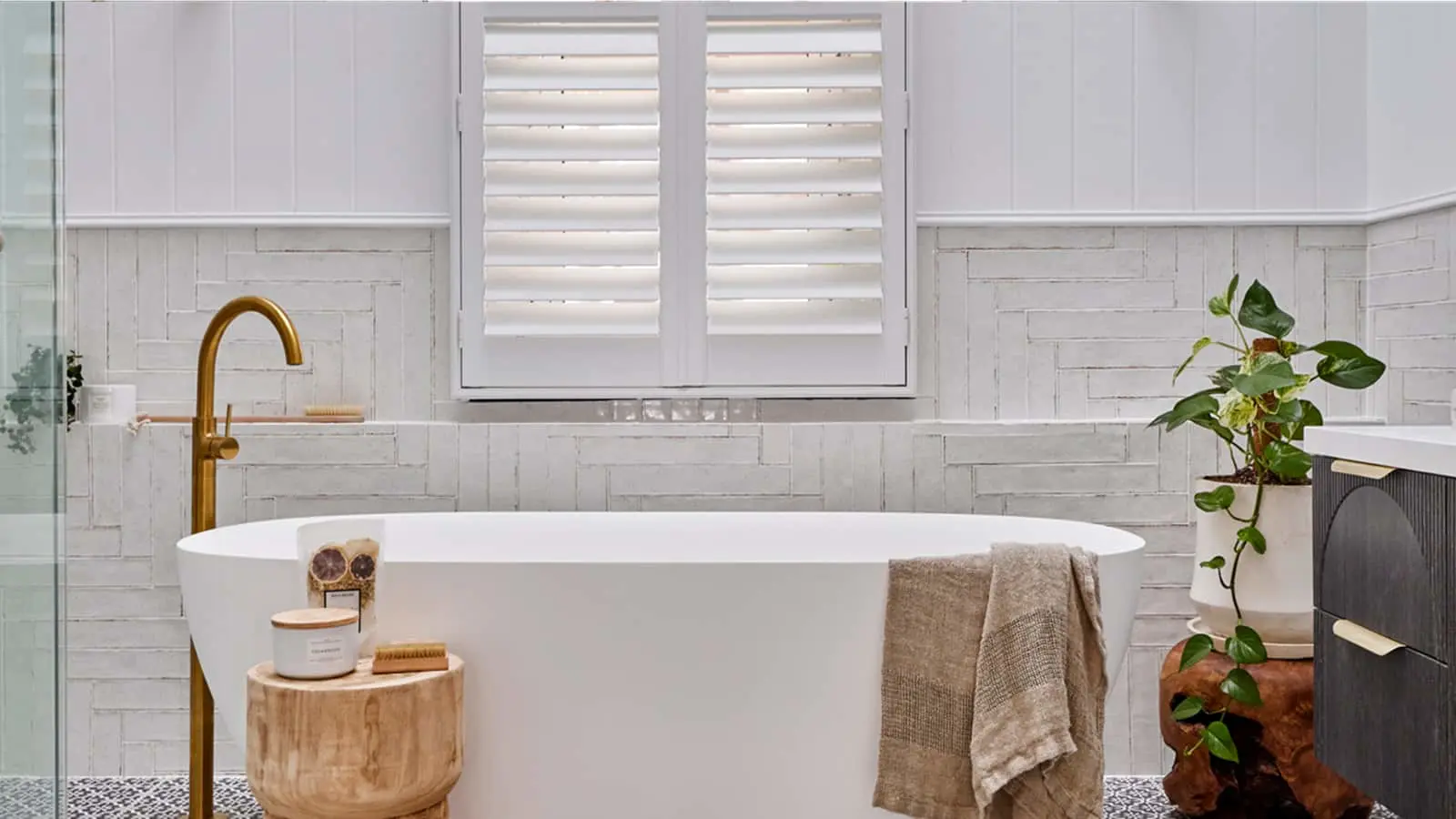 Modern white bathroom with freestanding tub, brass fixtures, wooden stool, white shutters, and potted plant.