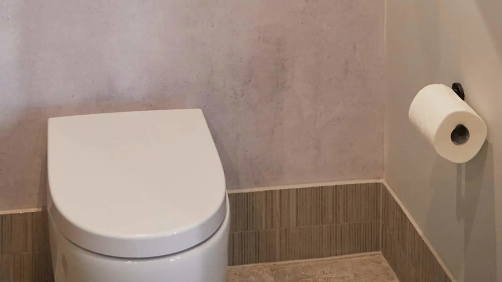 White toilet with closed lid in bathroom with gray walls, wooden paneling at bottom, and toilet paper roll mounted on wall.