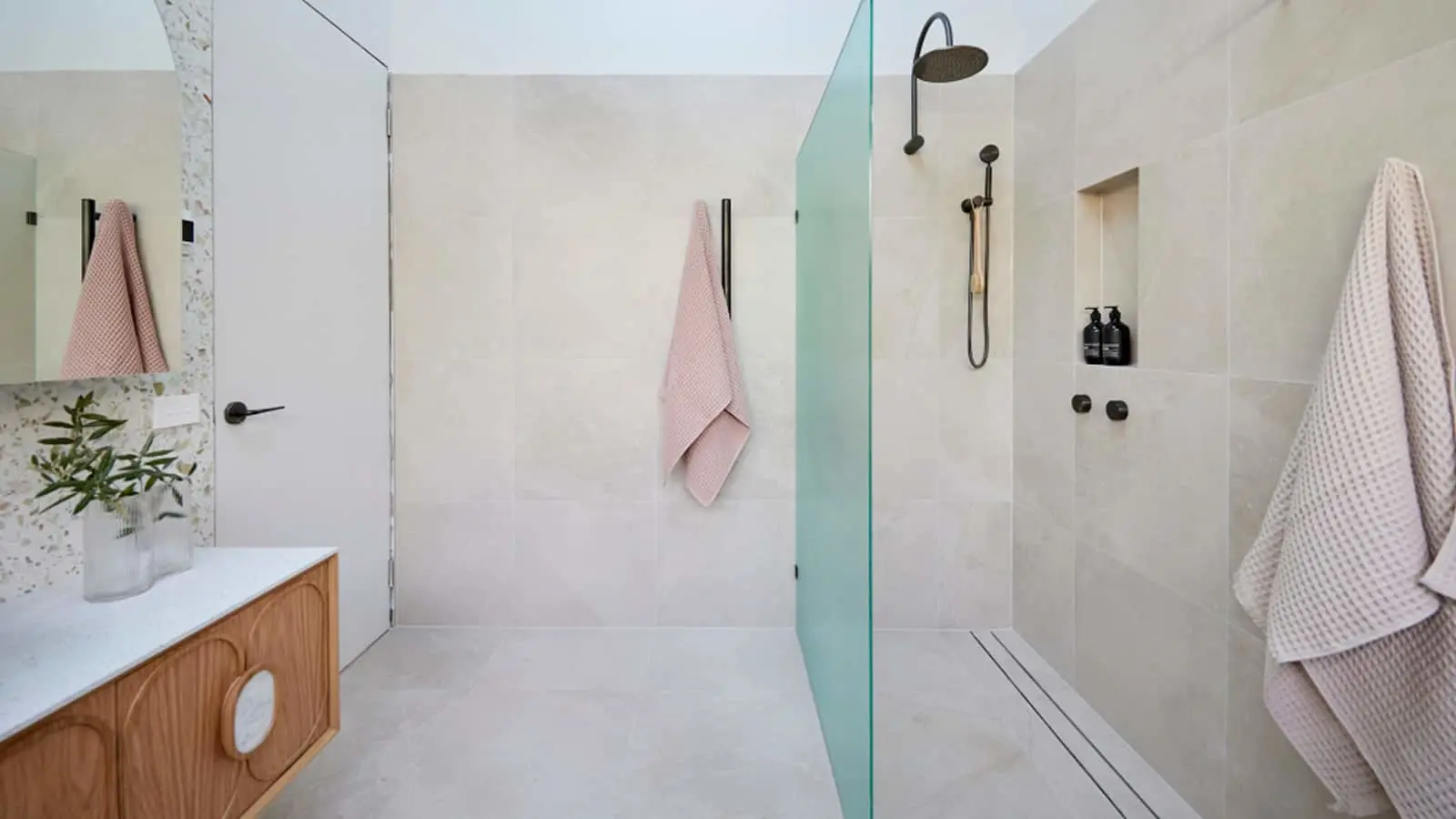 Modern bathroom with light walls, wooden vanity, glass shower partition, pink and white towels, and rainfall showerhead.