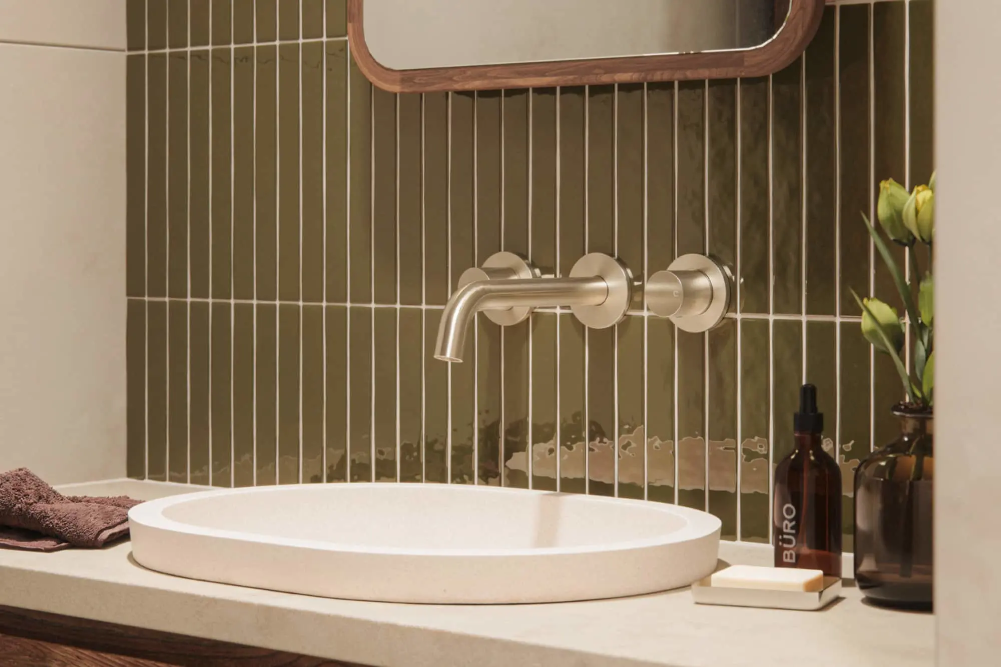 Modern bathroom sink with green vertical tile backsplash, wall-mounted faucet, wooden mirror, and decorative bottles.