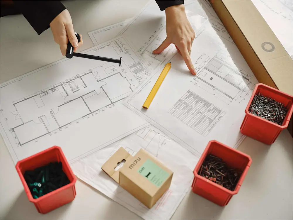 Hands reviewing architectural blueprints with measuring tool, pencil, and red containers of nails and fasteners on desk.