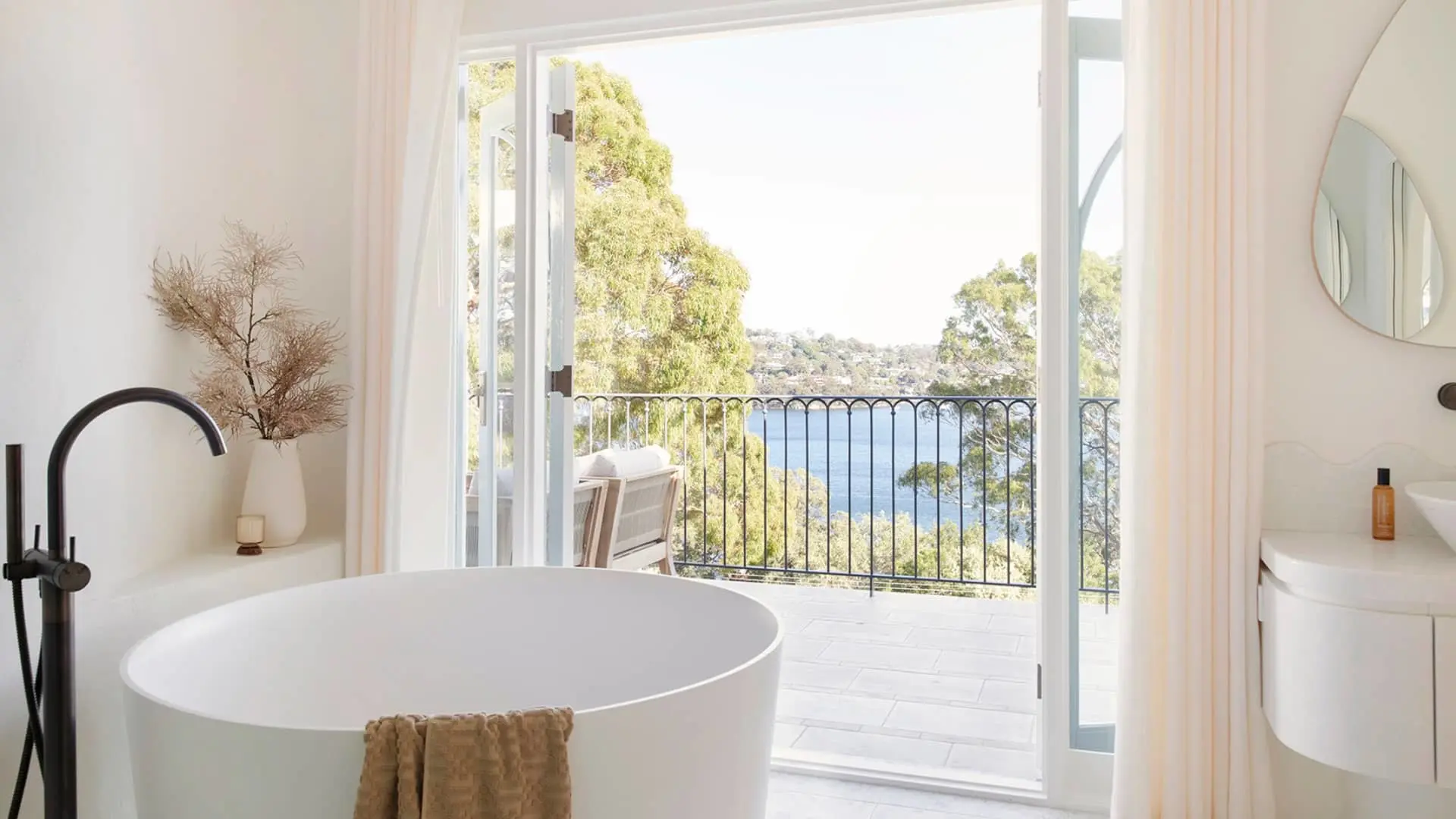 Modern bathroom with white freestanding tub and open doors revealing balcony with water view through trees.