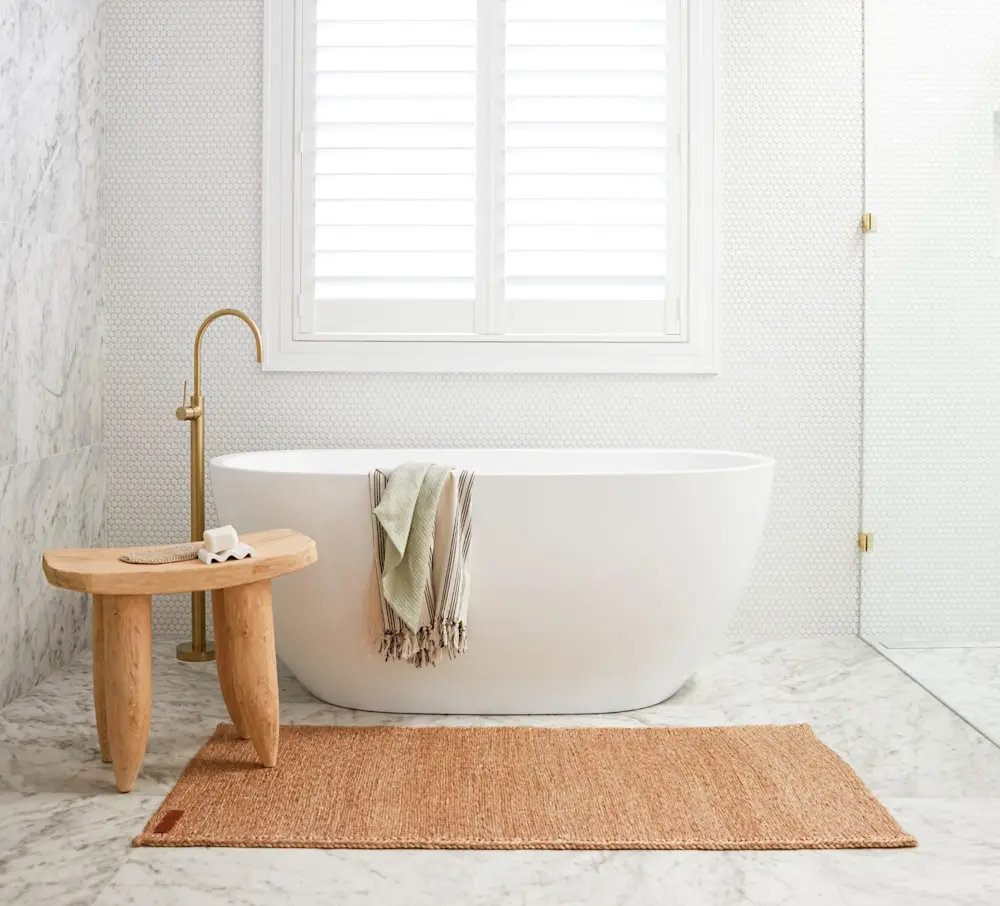 Modern white bathroom with freestanding tub, wooden stool, gold faucet, and natural fiber rug beneath window with shutters.