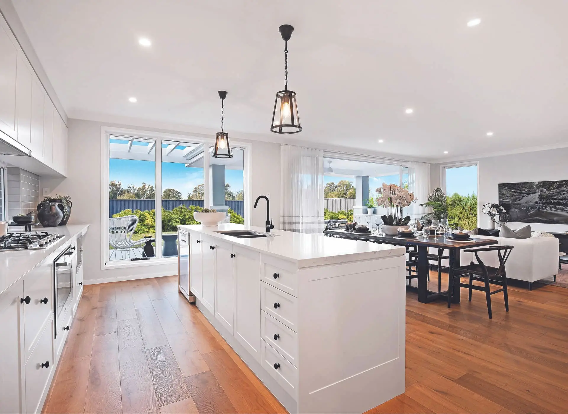 Modern white kitchen with island, wooden floors, pendant lights, and large windows overlooking garden and dining area.