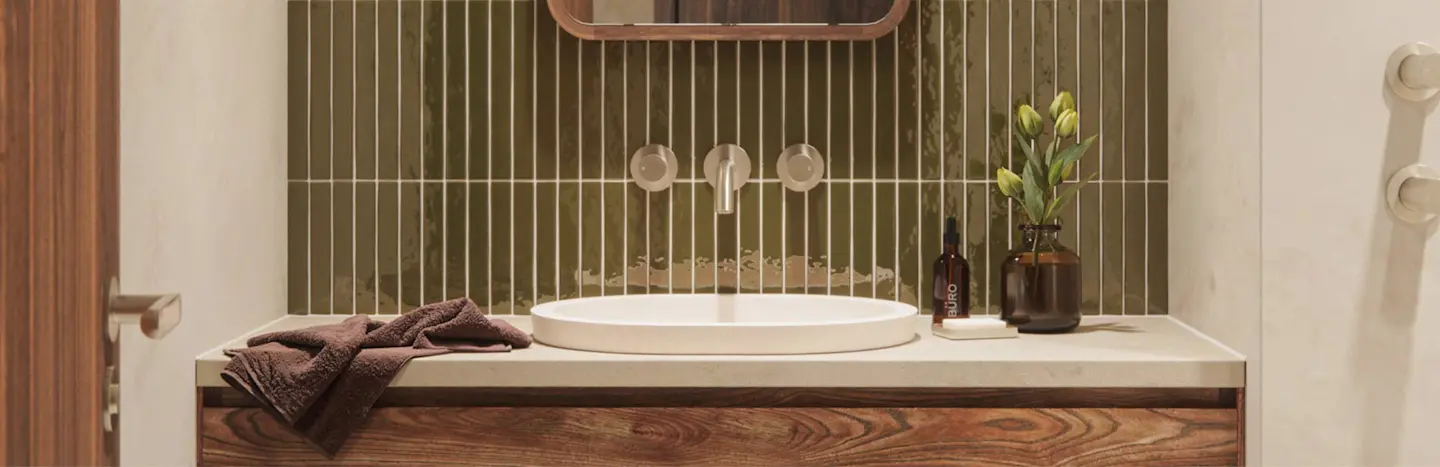 Modern bathroom with olive green tile backsplash, white sink, wooden vanity, brown towel, and small vase of yellow flowers.