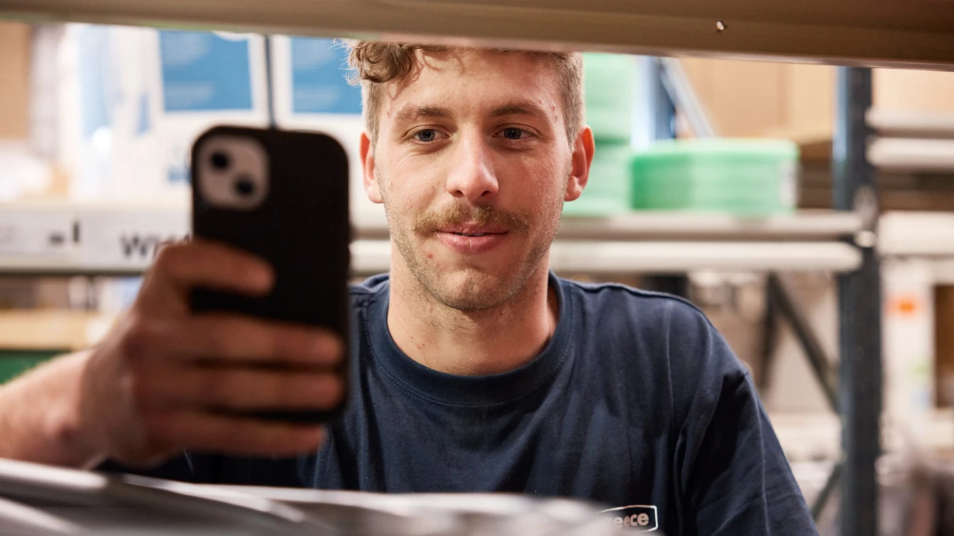 Person in blue shirt taking a selfie with black smartphone in what appears to be a workshop or industrial setting.