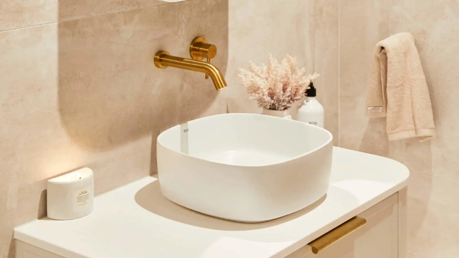 Modern bathroom with white vessel sink, gold wall-mounted faucet, beige walls, and decorative coral plant.