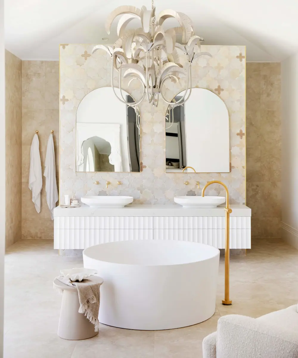 Elegant bathroom with white freestanding tub, dual vessel sinks, arched mirrors, and cream tile walls under a decorative chandelier.