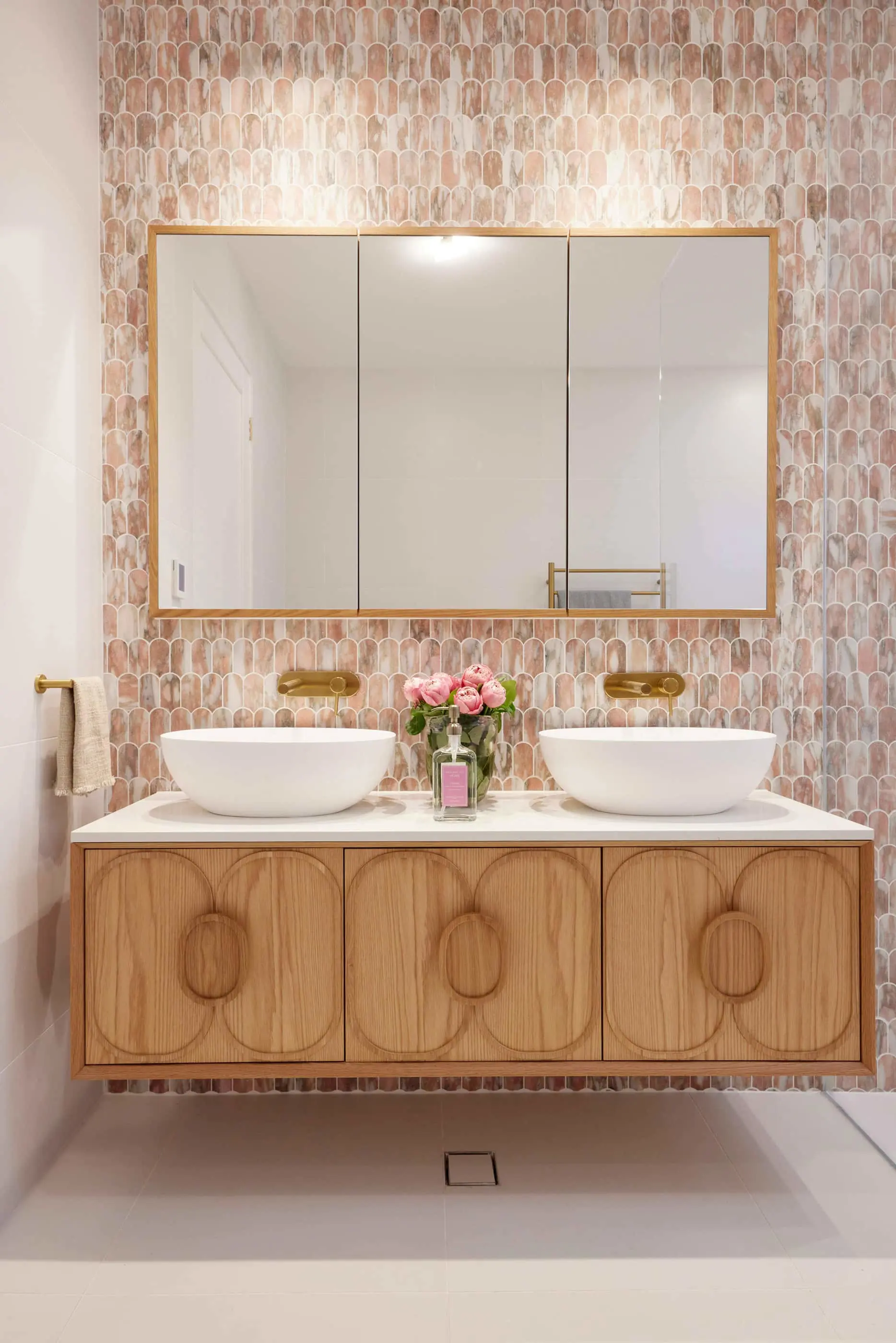 Modern bathroom with pink tile wall, wooden vanity with two white vessel sinks, gold fixtures, and pink roses centerpiece.