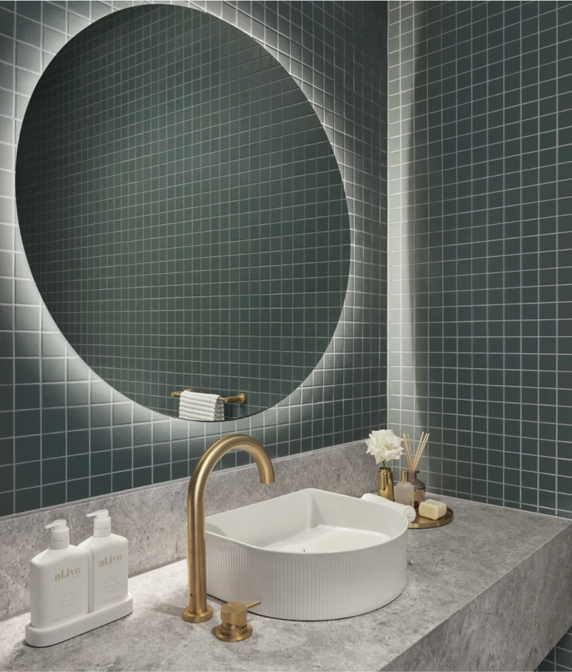 Modern bathroom with green tile walls, round backlit mirror, marble countertop, brass faucet, and white vessel sink.