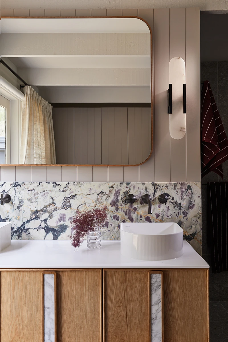 Modern bathroom with rounded mirror, marble backsplash, white sink on wooden vanity, and wall sconce light fixture.