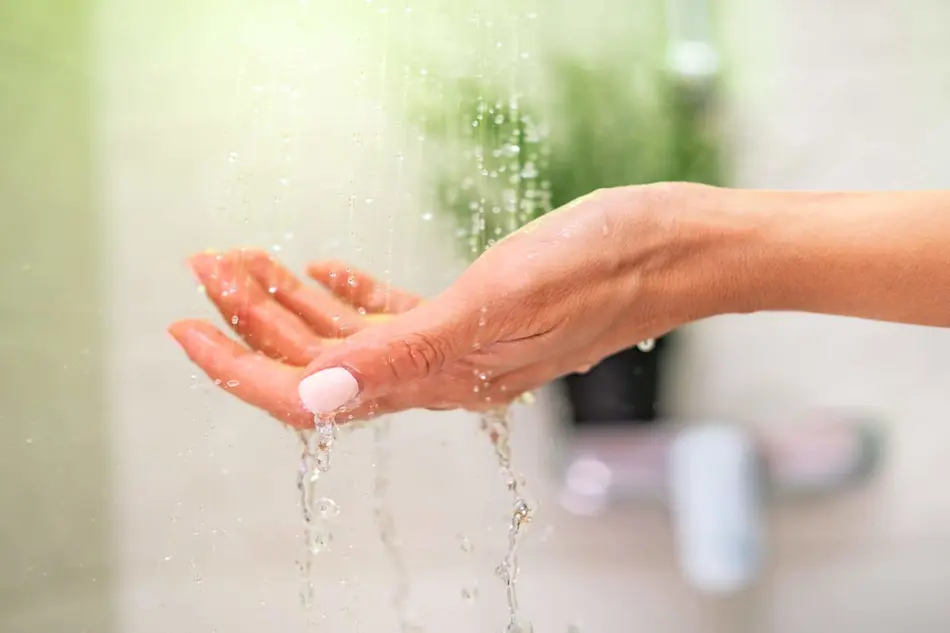 Hand with light pink nail polish catching water droplets in a bathroom shower with blurred green background.
