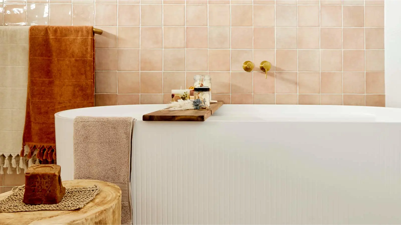 Spa-like bathroom with white bathtub, wooden bath tray, rust-colored towel, and beige tile walls with gold fixtures.