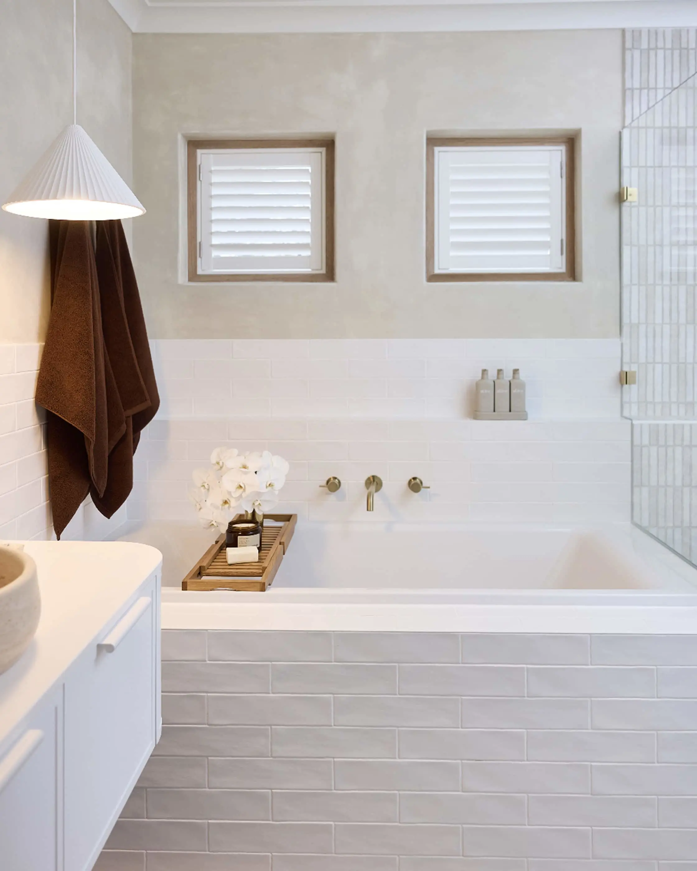 Modern white bathroom with bathtub, wooden tray, brown towel, pendant light, and two small windows with shutters.