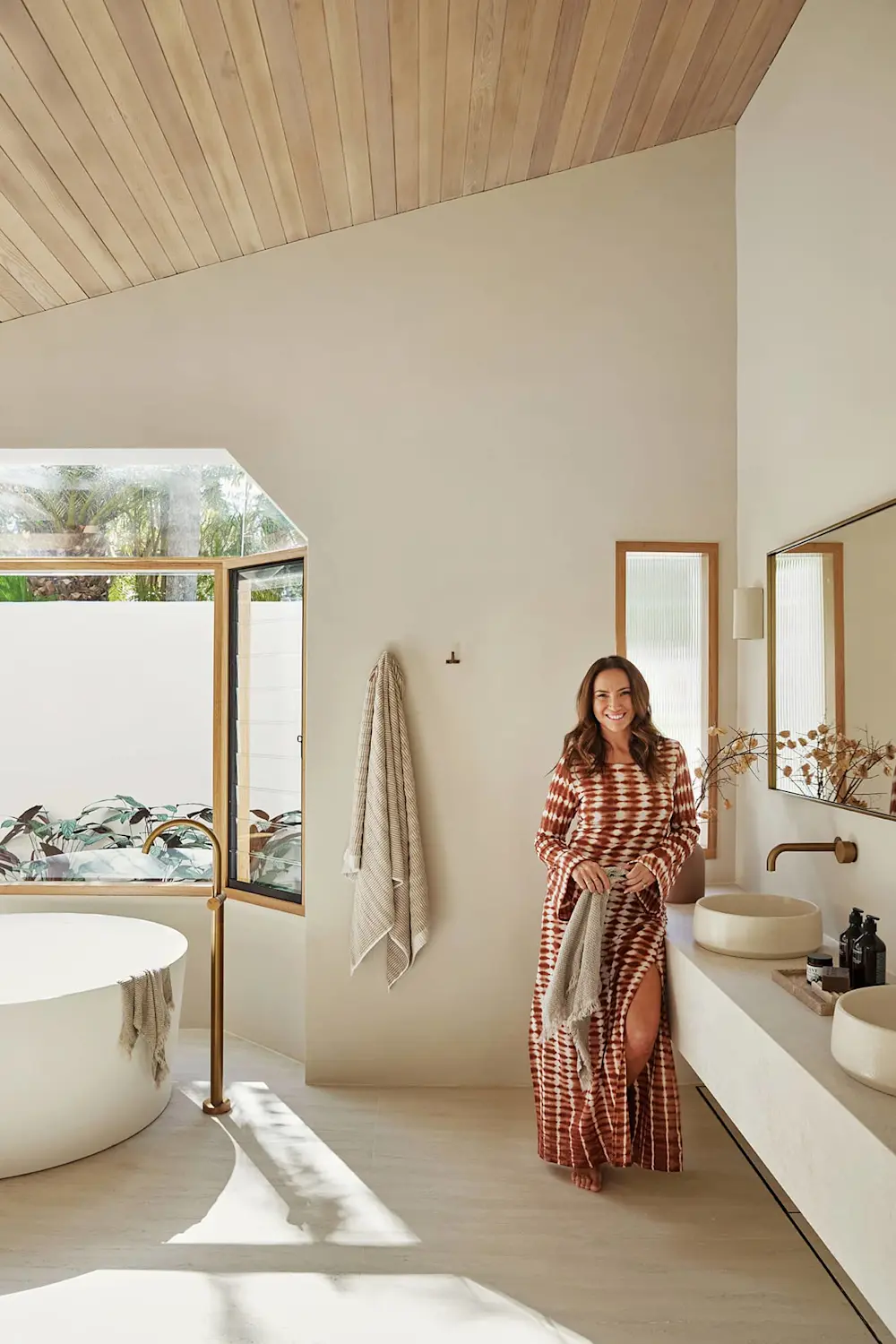 Modern bathroom with wooden ceiling, white bathtub, double sinks, and large windows overlooking greenery.