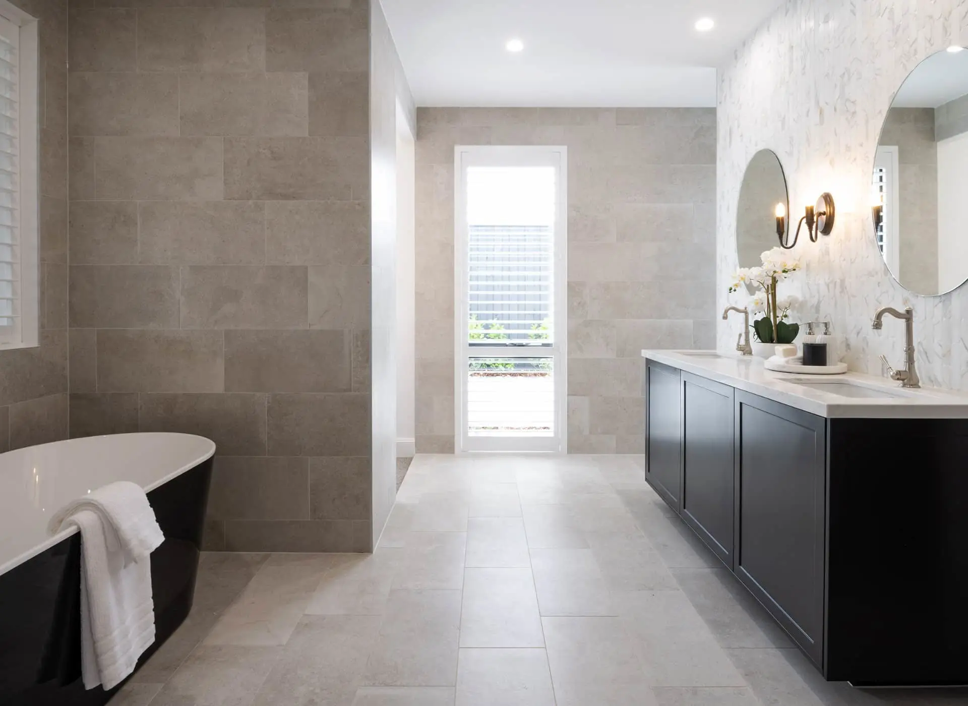 Modern bathroom with stone tile walls, freestanding black tub, navy vanity, round mirror, and tall window with blinds.