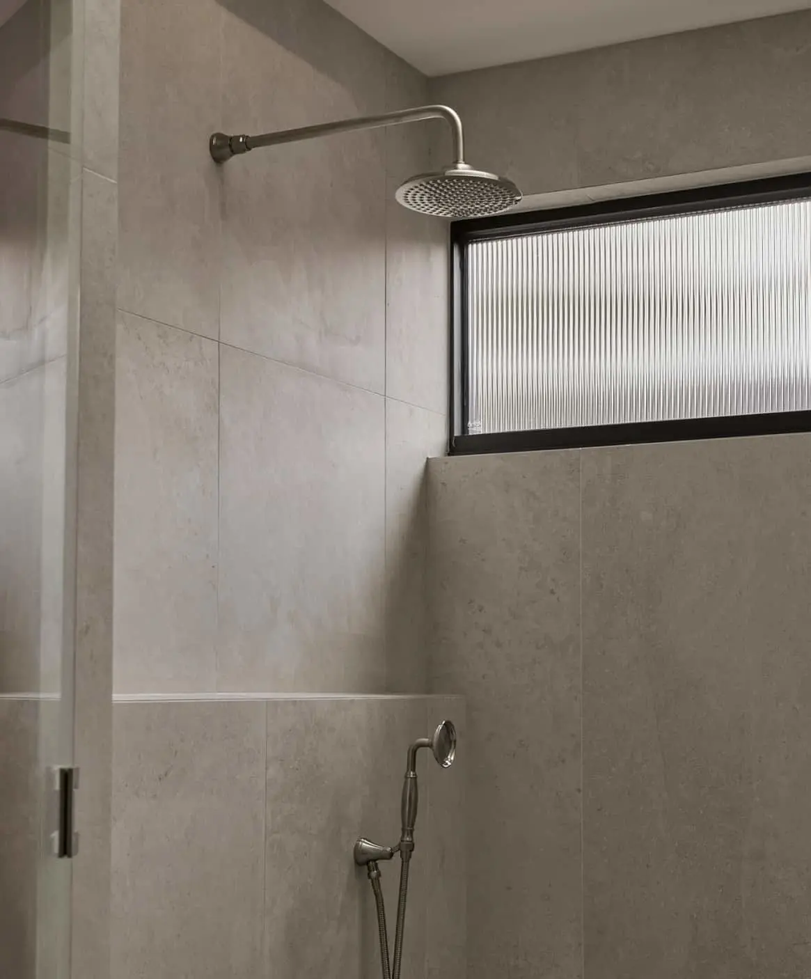 Modern shower with beige stone tiles featuring overhead rainfall showerhead and handheld sprayer, with frosted window.