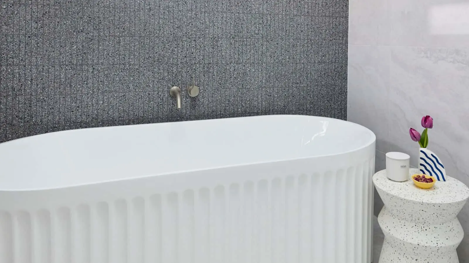 Modern bathroom with white freestanding tub, textured gray wall, and small side table holding pink tulips and a candle.
