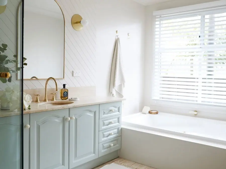 Bright bathroom with mint green vanity, gold-framed mirror, brass fixtures, white bathtub, and window with blinds.