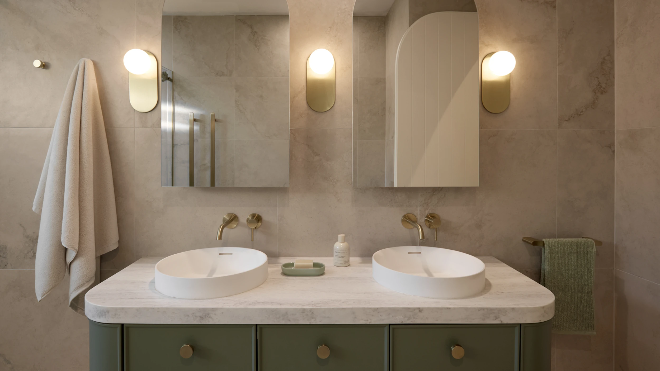 Modern bathroom with dual white sinks on marble countertop, green vanity, oval mirrors, and brass fixtures.