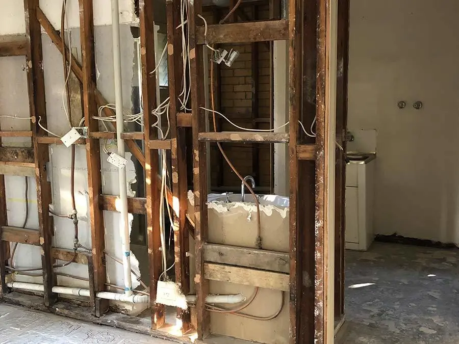 Exposed wooden wall studs with electrical wiring in a home renovation, showing plumbing fixtures and partial drywall removal.