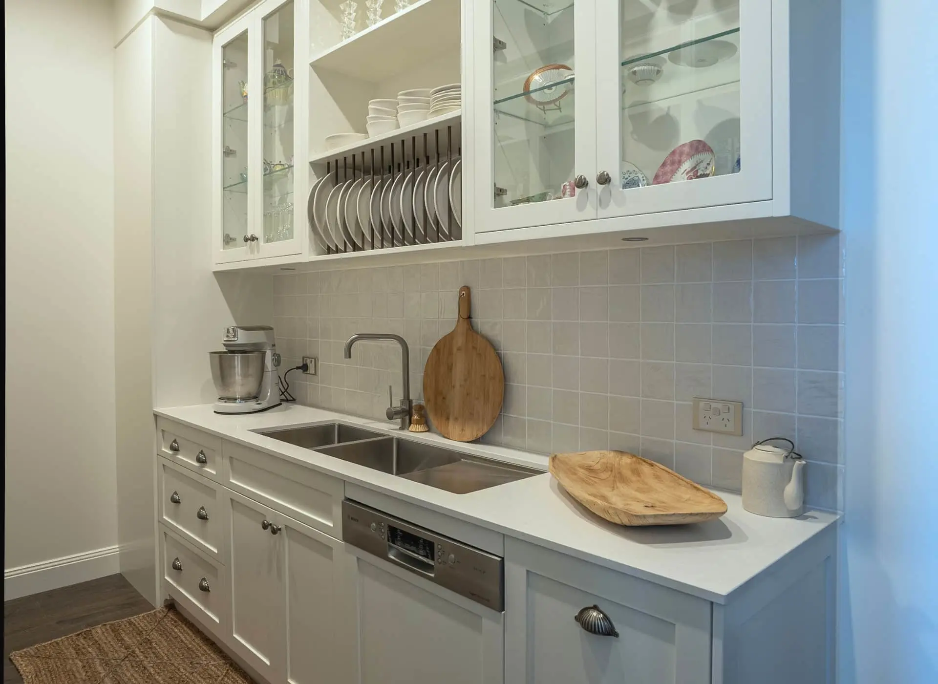 White kitchen with glass cabinets displaying dishes, sink with wooden cutting board, mixer, and dishwasher below.