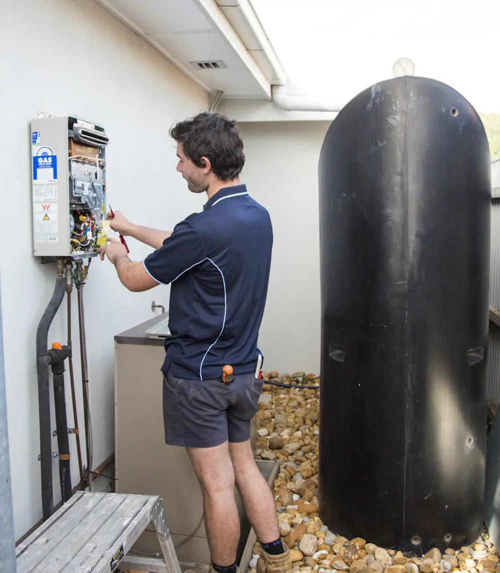 Technician in blue polo shirt repairing a gas water heater system next to a large black water tank.