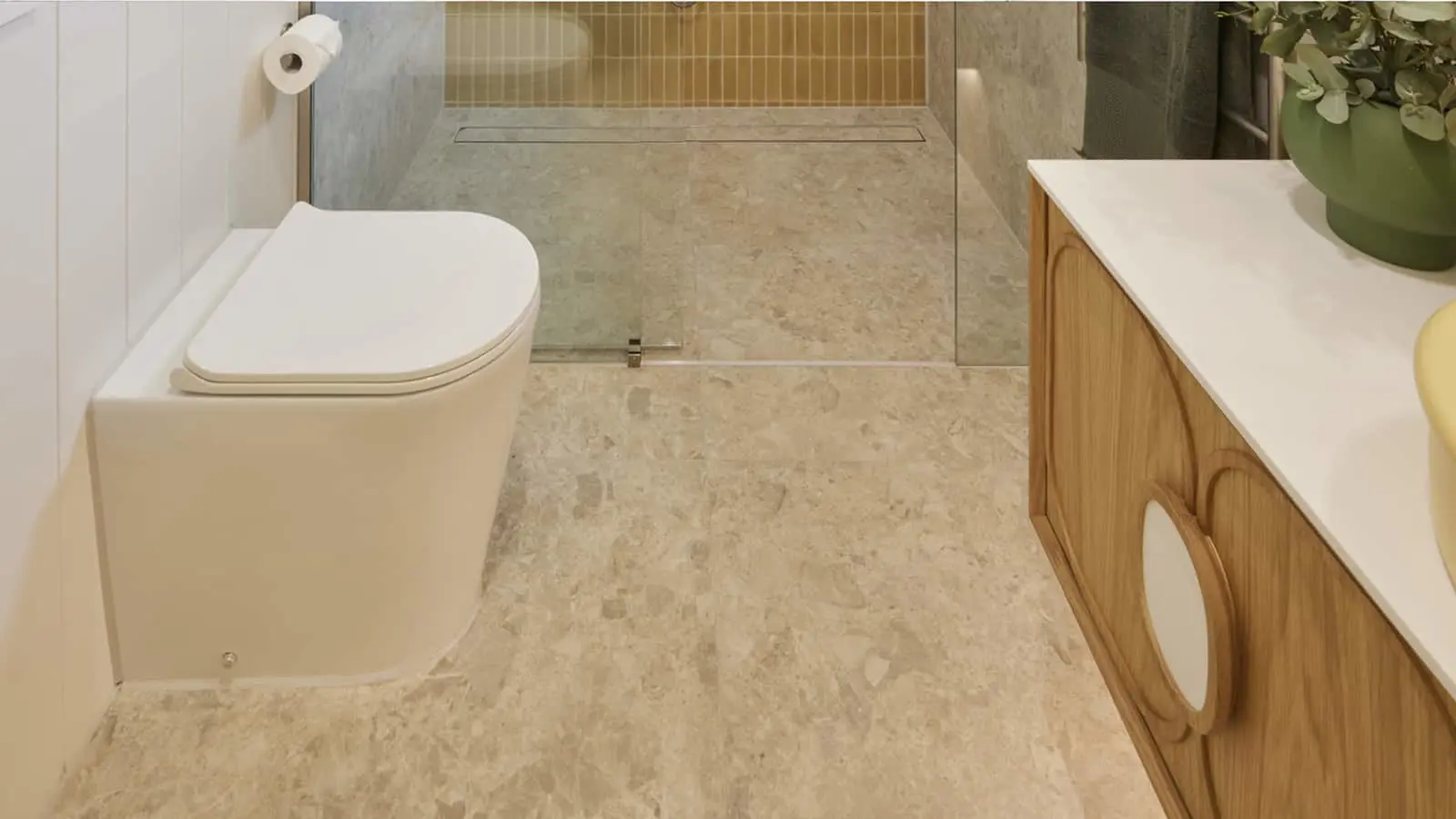 Modern bathroom with white toilet, wooden vanity, stone tile floor, and glass shower enclosure with linear drain.