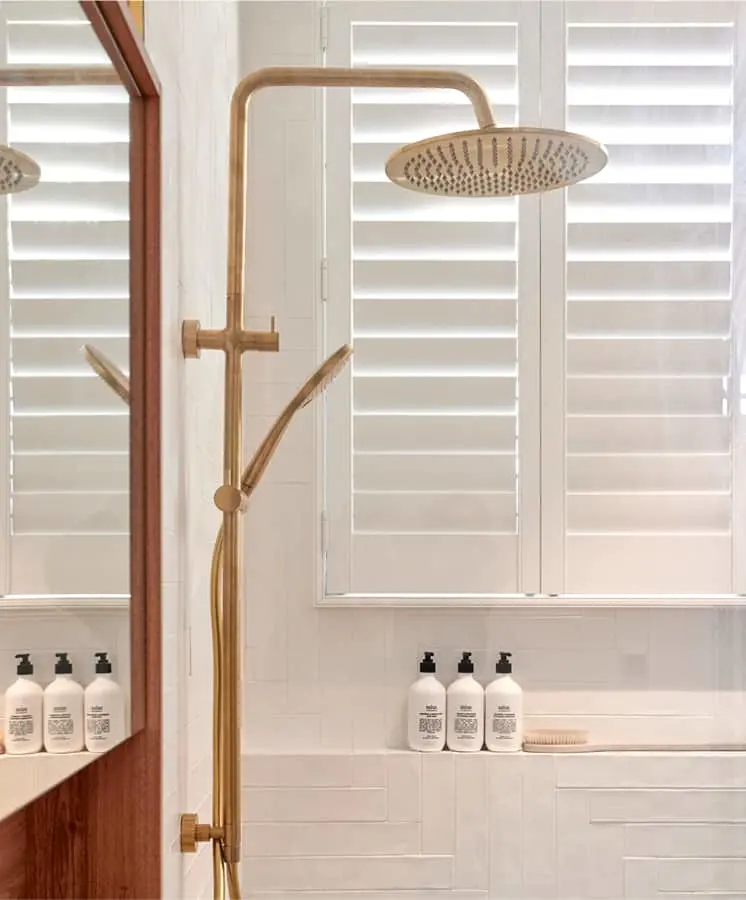 Modern bathroom with brass rainfall shower fixture, white shuttered windows, and toiletry bottles on a shelf.