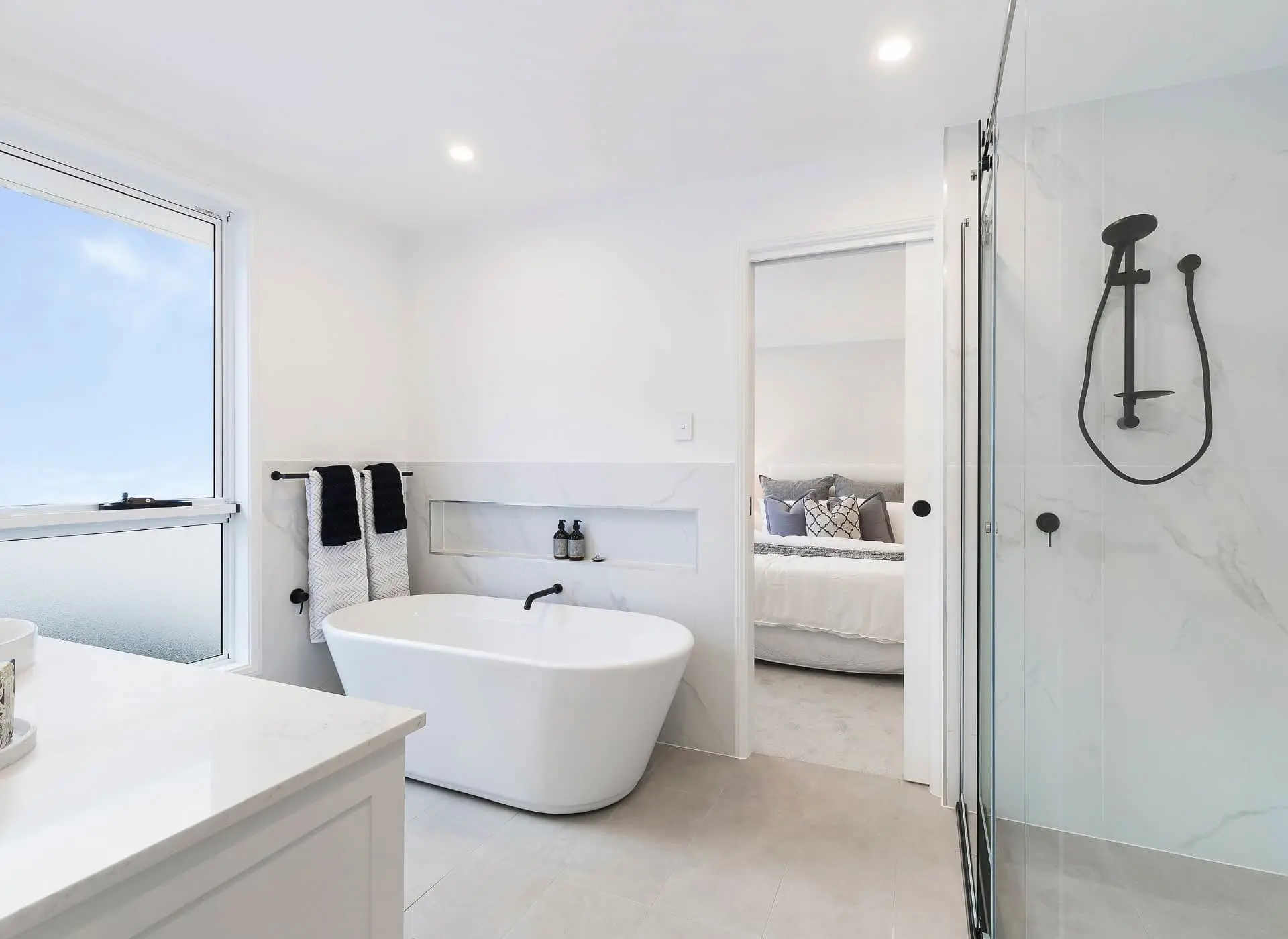 Modern white bathroom with freestanding tub, glass shower, and view into bedroom with gray pillows.