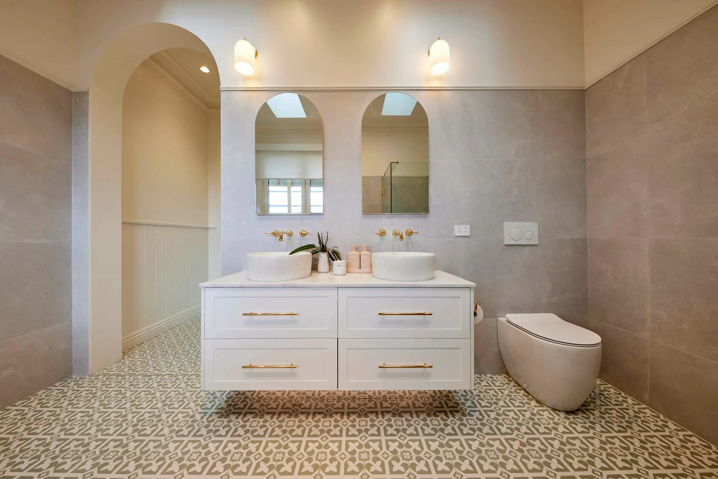 Elegant bathroom with white vanity, dual vessel sinks, gold fixtures, patterned floor tiles, and arched doorway.