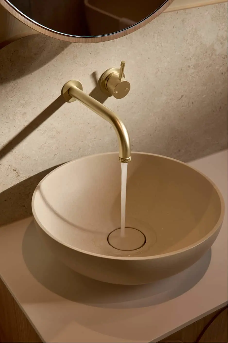 Round beige basin on a countertop with water running from a wall-mounted brushed gold faucet and tap handle.