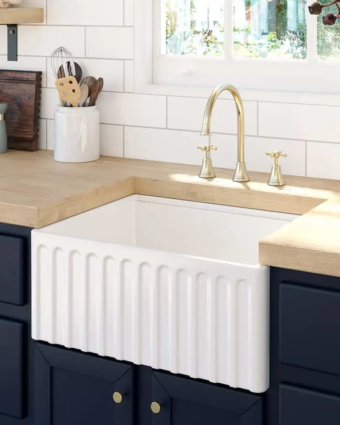 White farmhouse sink with brass faucet in kitchen with wooden countertops, navy cabinets, and utensil holder.
