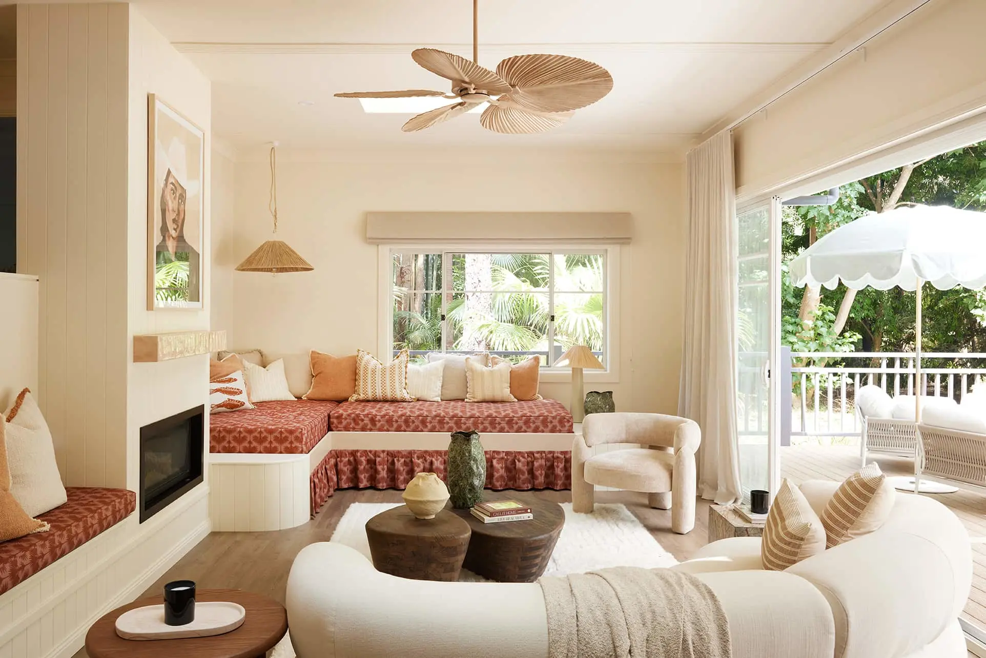 Bright coastal living room with palm leaf ceiling fan, terracotta daybed, white sofa, and patio doors opening to garden.