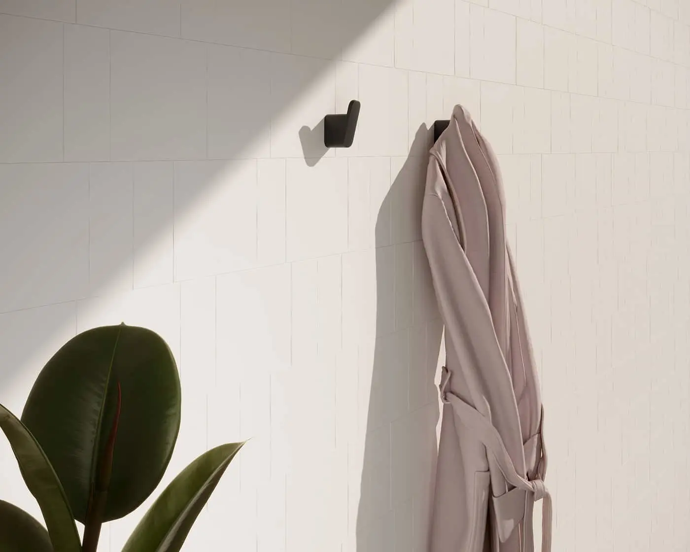 Light pink bathrobe hanging on black wall hook with rubber plant leaves visible in corner against white tiled wall.