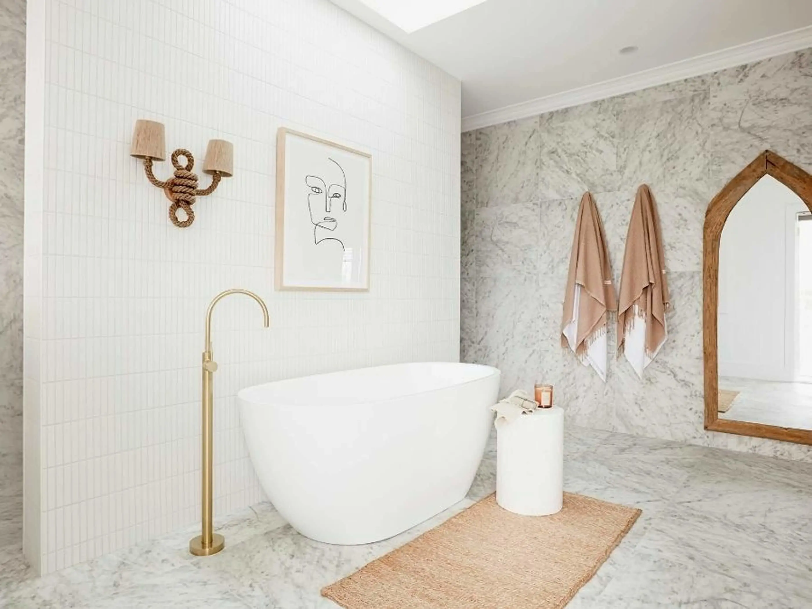 Modern bathroom with white freestanding tub, marble walls, rope sconce, line art, and peach towels hanging beside wooden mirror.