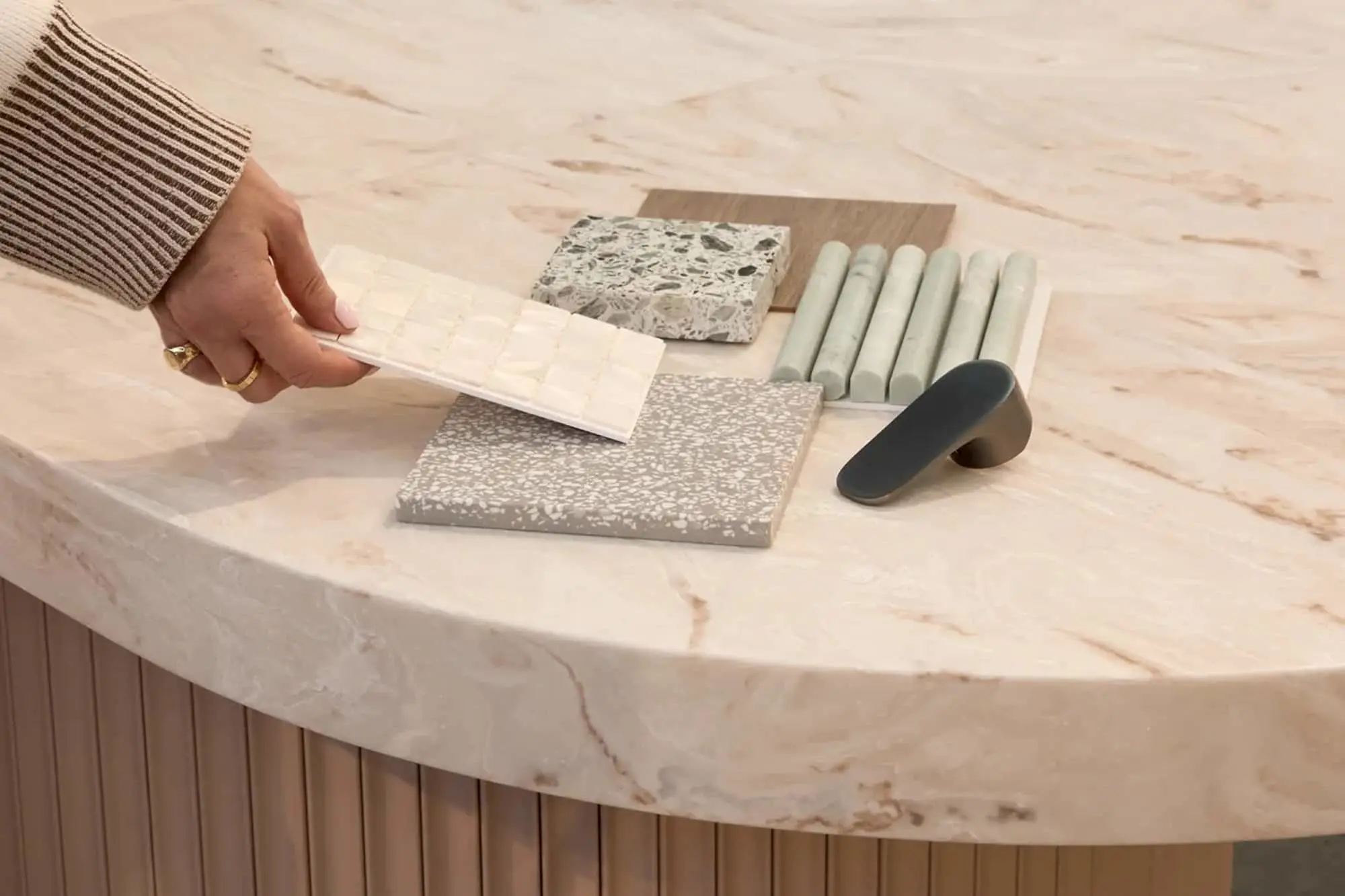 A hand holding a white mosaic tile sample over a marble table with terrazzo, stone, and hardware samples.